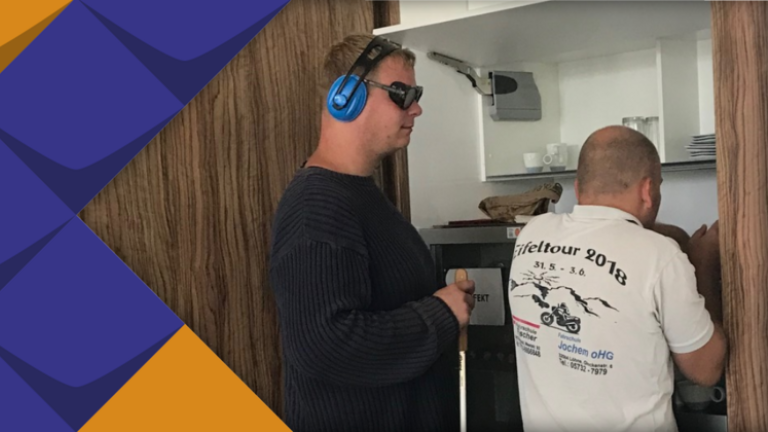 Zwei Männer an einer Kaffeemaschine. Einer davon mit dunkler Brille, Gehörschutz und Blindenstock.