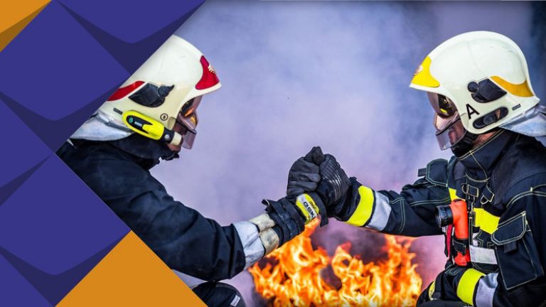 Zwei Feuerwehrmitarbeiter greifen sich an die Hände. Im Hintergrund lodert ein Feuer.